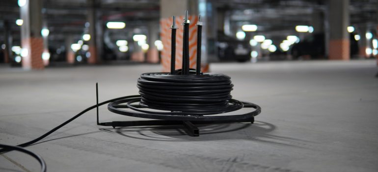 A coiled black heavy-duty cable on a metal dispenser stand sitting on the floor of a dimly lit underground parking garage.